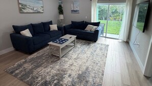 Una sala de estar con dos sofás oscuros, una mesa de centro, un ventilador de techo y un ventanal con vistas a zonas verdes.