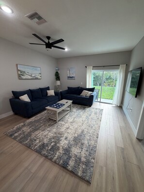 A living room with two dark sofas, a coffee table, a ceiling fan, and a large window with a view of greenery. - Gorgeous Coastal Townhome - 8 min from Boca Grande, Close to Beaches & Shops (Port Charlotte)