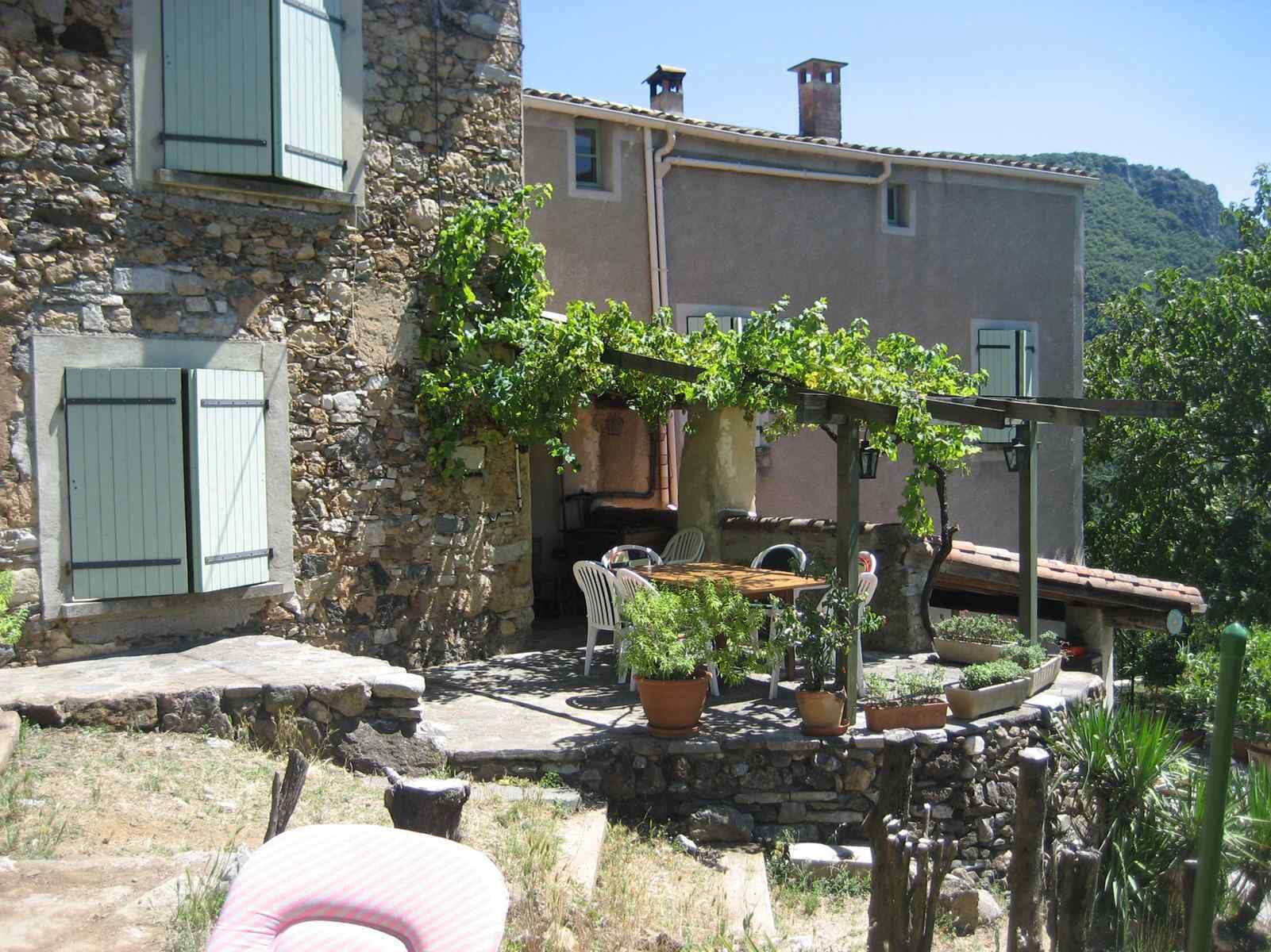 Ein Steinhaus mit Garten, der mit Kübelpflanzen und einer mit Wein bewachsenen Pergola gestaltet ist.