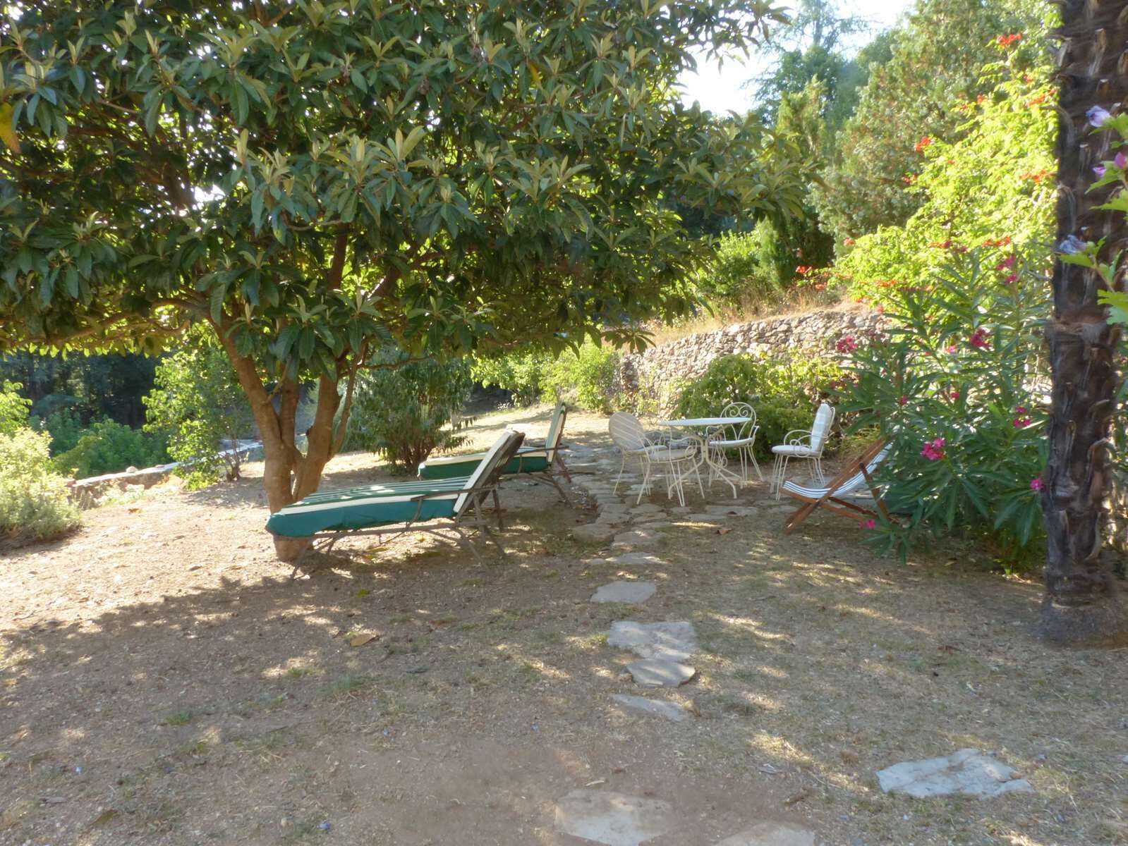 Ein Sitzbereich im Freien mit einer Steinsäule, einem Baum, der Schatten spendet, und Blick auf einen Garten.