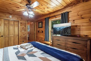 Une chambre dans un chalet en bois, équipée d’un lit, d’un ventilateur de plafond, d’une télévision et d’une fenêtre donnant sur les montagnes.