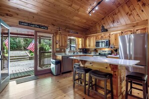 Une cuisine rustique aménagée dans une cabane en bois, avec un îlot central, des appareils en acier inoxydable et une vue sur l’extérieur.