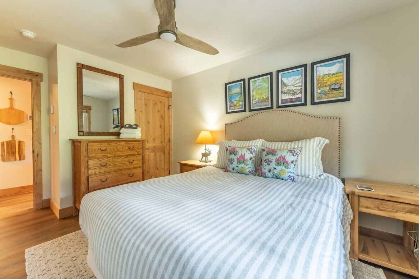 A bedroom with a bed, wooden dresser, nightstand, lamp, ceiling fan, and framed pictures on the wall.