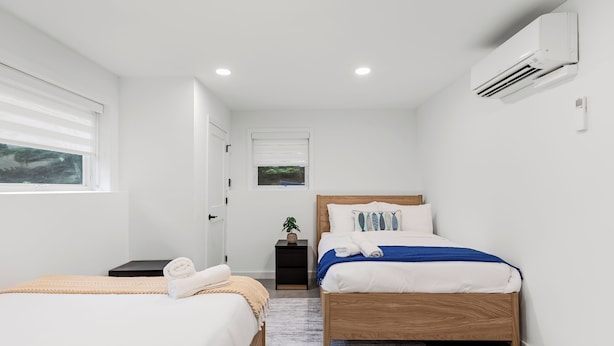 Un dormitorio moderno con dos camas, una mesita de noche pequeña, una planta en maceta y un equipo de aire acondicionado.