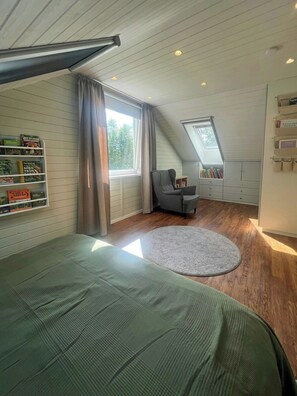 Une chambre avec un plafond en pente, un lit, une chaise, une fenêtre et une bibliothèque.