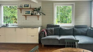 A modern living room with a grey sofa, wooden dining table, and a kitchen area with white cabinets and a window overlooking greenery. - Lakeside cottage with private beach in Sörmland (Stavsjö)