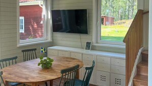 A dining area with a wooden table and chairs, a television mounted above a bench, and a view of a red building through a window. - Lakeside cottage with private beach in Sörmland (Stavsjö)