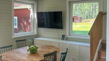 Un coin repas avec une table et des chaises en bois, un téléviseur fixé au-dessus d’un banc, et une vue sur un bâtiment rouge par la fenêtre.