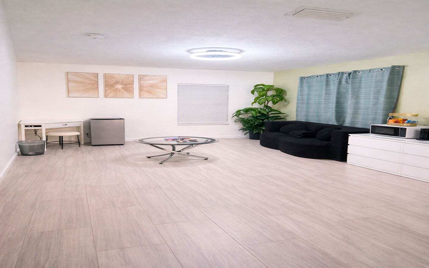 Un amplio salón con una mesa redonda de vidrio, un sofá negro, un armario blanco y una planta en maceta.