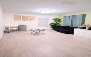 Un amplio salón con una mesa redonda de vidrio, un sofá negro, un armario blanco y una planta en maceta.