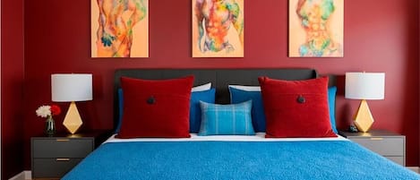 Un dormitorio con cama, almohadas rojas y azules, dos mesitas de noche con lámparas y tres cuadros abstractos en la pared.