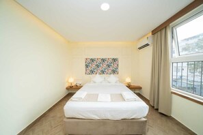 A hotel room with a bed, two bedside tables with lamps, a window with curtains, and a wall-mounted air conditioner. - The Clock Tower Inn (Bengaluru)