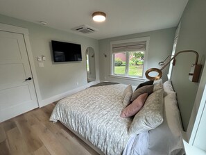 Ein Schlafzimmer mit einem Bett, einem an der Wand montierten Fernseher, einem Fenster mit Jalousien und einer Deckenleuchte.