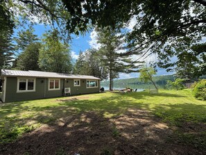 A cabin with a metal roof, surrounded by a grassy yard and trees, overlooking a lake. - Luxury Lakefront | Golf Sim & Theatre | Dock (Westport)
