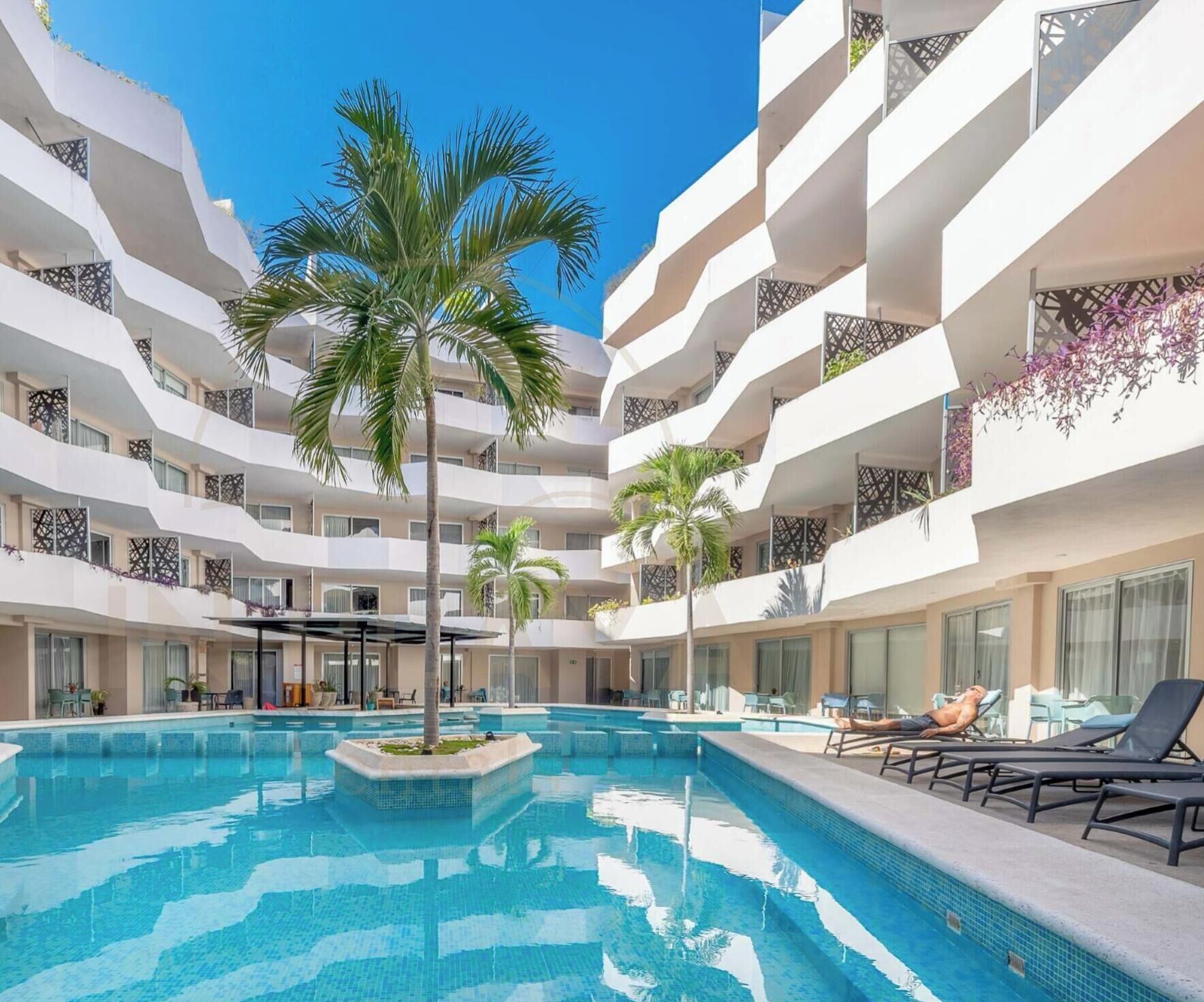 Un hôtel à plusieurs étages doté d’une piscine centrale, entourée de chaises longues et de palmiers.