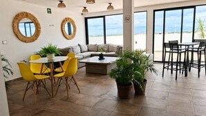 Uma sala ampla com sofá, poltronas amarelas, mesa de centro e grandes janelas com vista para o céu e a paisagem distante.