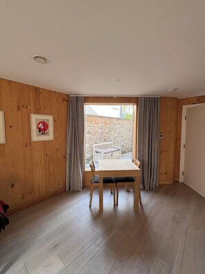 Ein Zimmer mit Holzboden, einem Tisch mit Stühlen, einem Fenster mit Vorhängen und einer Tür.