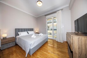 Un dormitorio con cama, piso de madera, televisor, balcón con cortinas y mesita de noche con lámpara.
