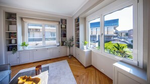 A modern living room with built-in bookshelves, a large window with a view of buildings, and a wooden floor. - Elenas Luxury Apartment/Panoramic Acropolis View (Athina)