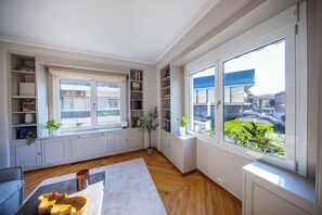 A modern living room with built-in bookshelves, a large window with a view of buildings, and a wooden floor. - Elenas Luxury Apartment/Panoramic Acropolis View (Athina)