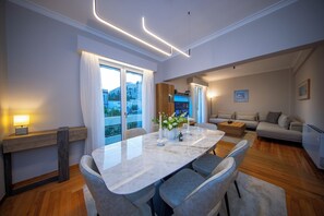 A modern living room with a dining area, featuring a marble table, grey upholstered chairs, a sofa, and a view of a cityscape through large windows. - Elenas Luxury Apartment/Panoramic Acropolis View (Athina)