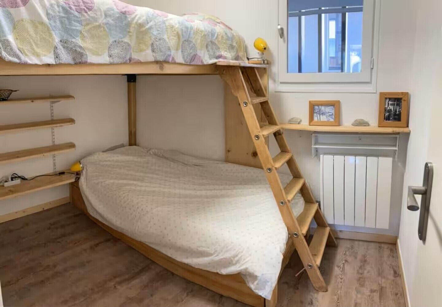 Ein Stockbett mit einer Holzleiter, einem kleinen Regal und einem Fenster mit Jalousien.