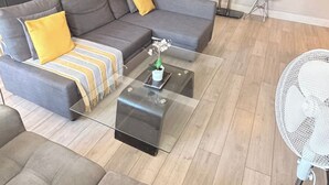 A modern living room with a sectional sofa, a glass coffee table, a small side table, and a white fan. - Marble Arch Elevator Ac - 3 Bedroom Apartment (London)