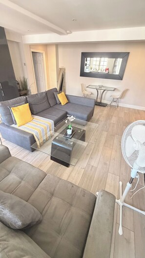 A modern living room with a sectional sofa, a glass coffee table, a small side table, and a white fan. - Marble Arch Elevator Ac - 3 Bedroom Apartment (London)