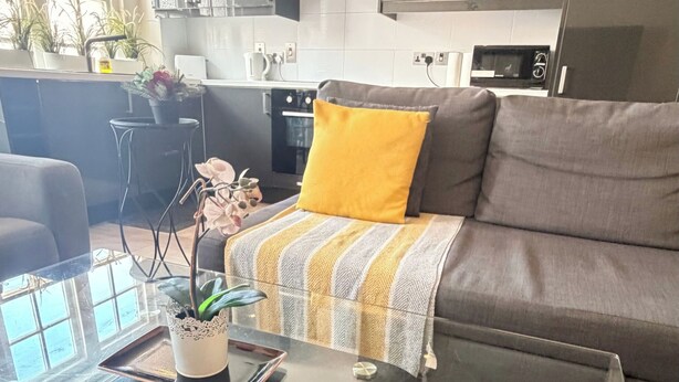 A modern living room with a glass coffee table, a sofa with a yellow pillow, and a kitchen in the background. - Marble Arch Elevator Ac - 3 Bedroom Apartment (London)