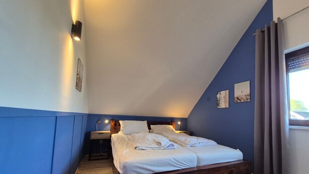 A bedroom with a wooden bed, a nightstand, a wall-mounted lamp, and a window with curtains. - Familienfreundliches Ferienhaus mit Garten im Hafendorf Muritz (Rechlin)