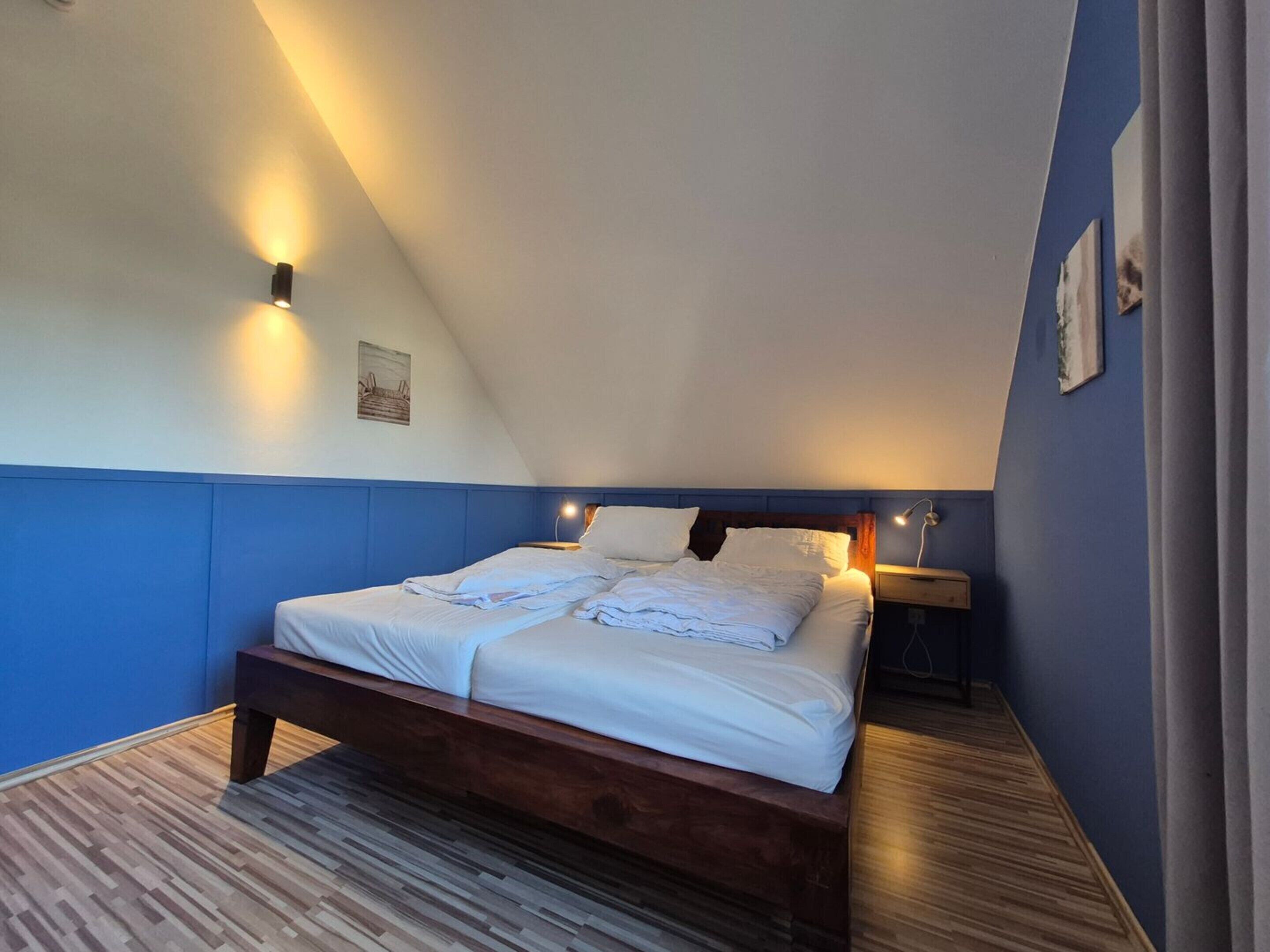 Une chambre avec un plafond en pente, un lit recouvert de draps blancs, une table de chevet avec une lampe et un plancher en bois.