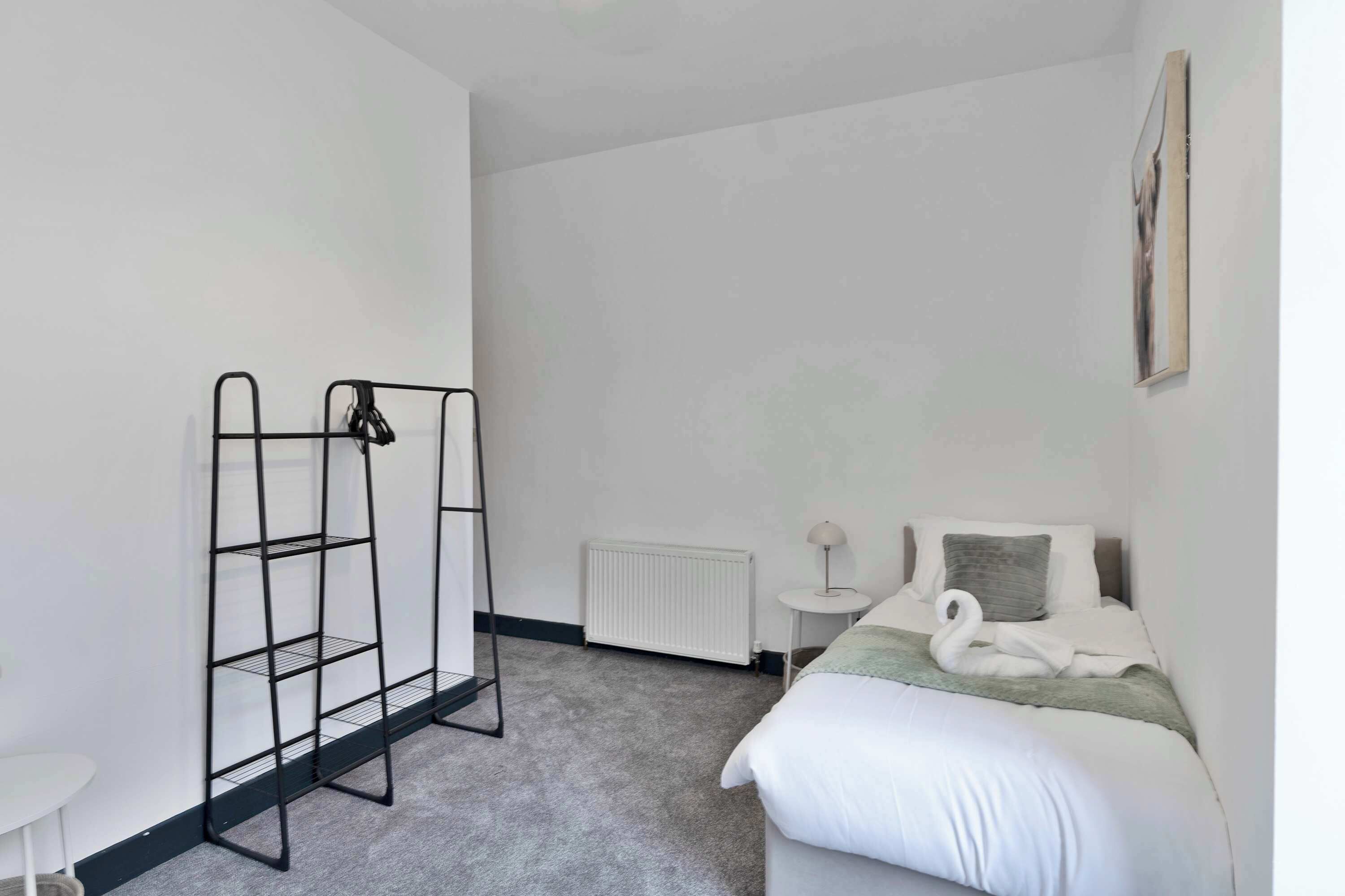 Une chambre avec un lit, un radiateur, une petite table et une étagère en métal noir.