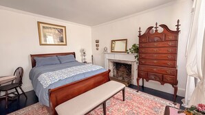 Un dormitorio con cama, banco, cómoda, chimenea y una silla.