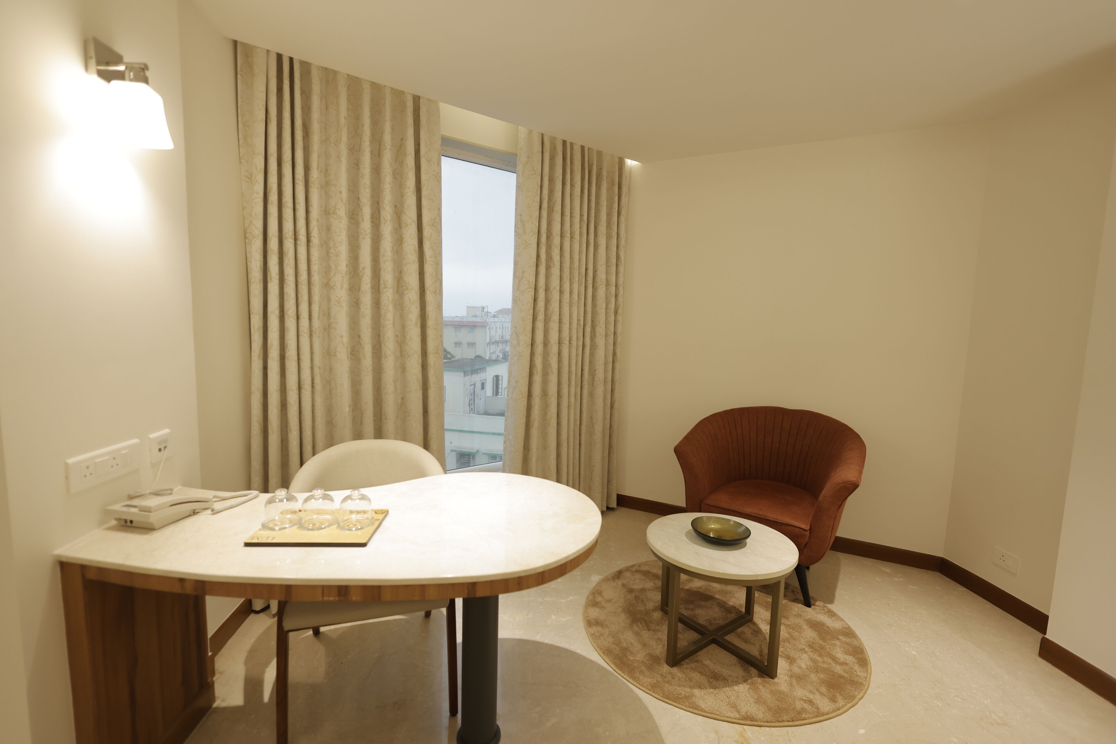 Habitación de hotel con una mesa de comedor, una mesita auxiliar, un sillón rojo y vista a la ciudad desde la ventana.