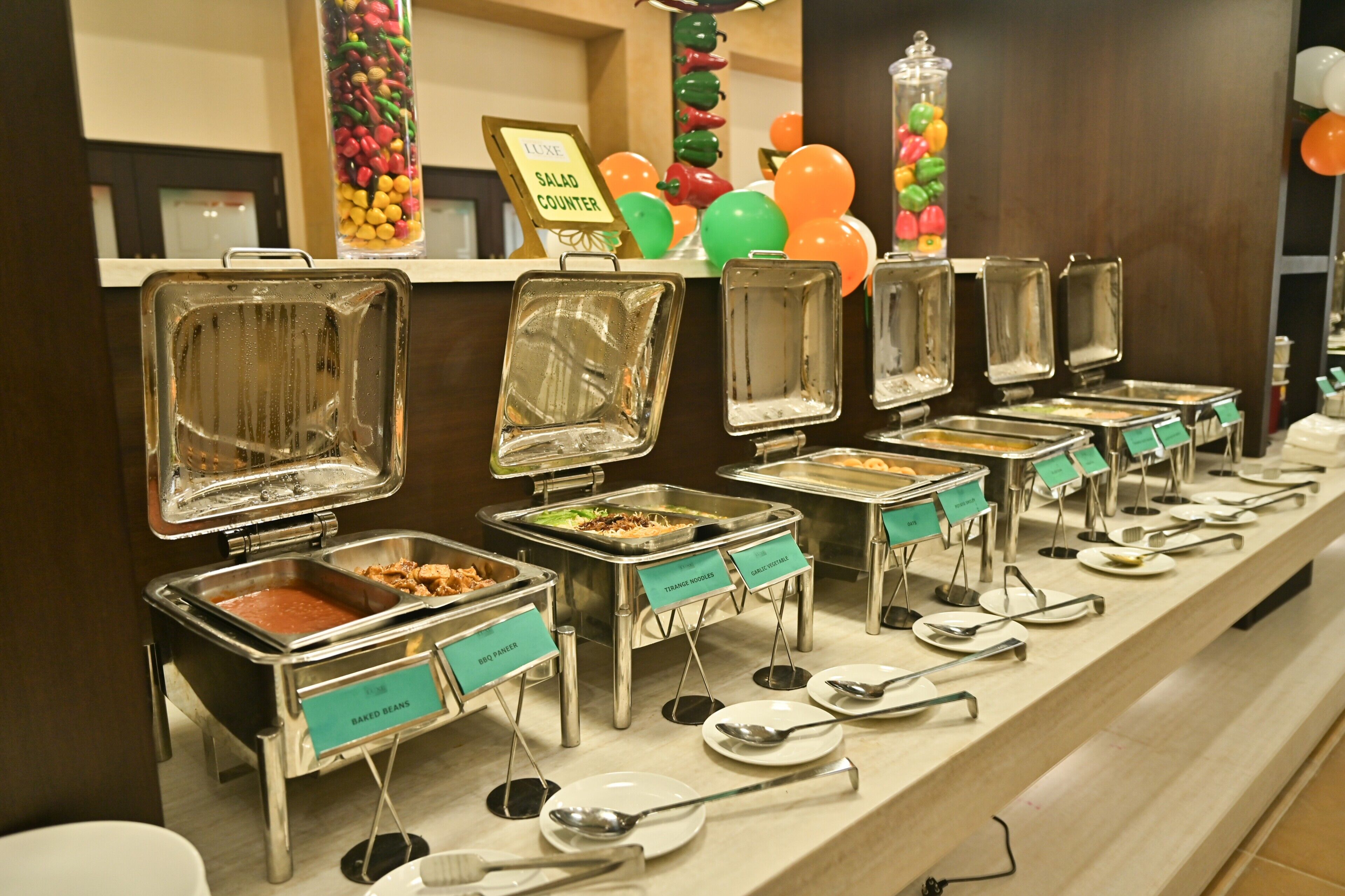 A buffet line with various food stations labeled with names like 'BBQ Paneer' and 'Baked Beans.'