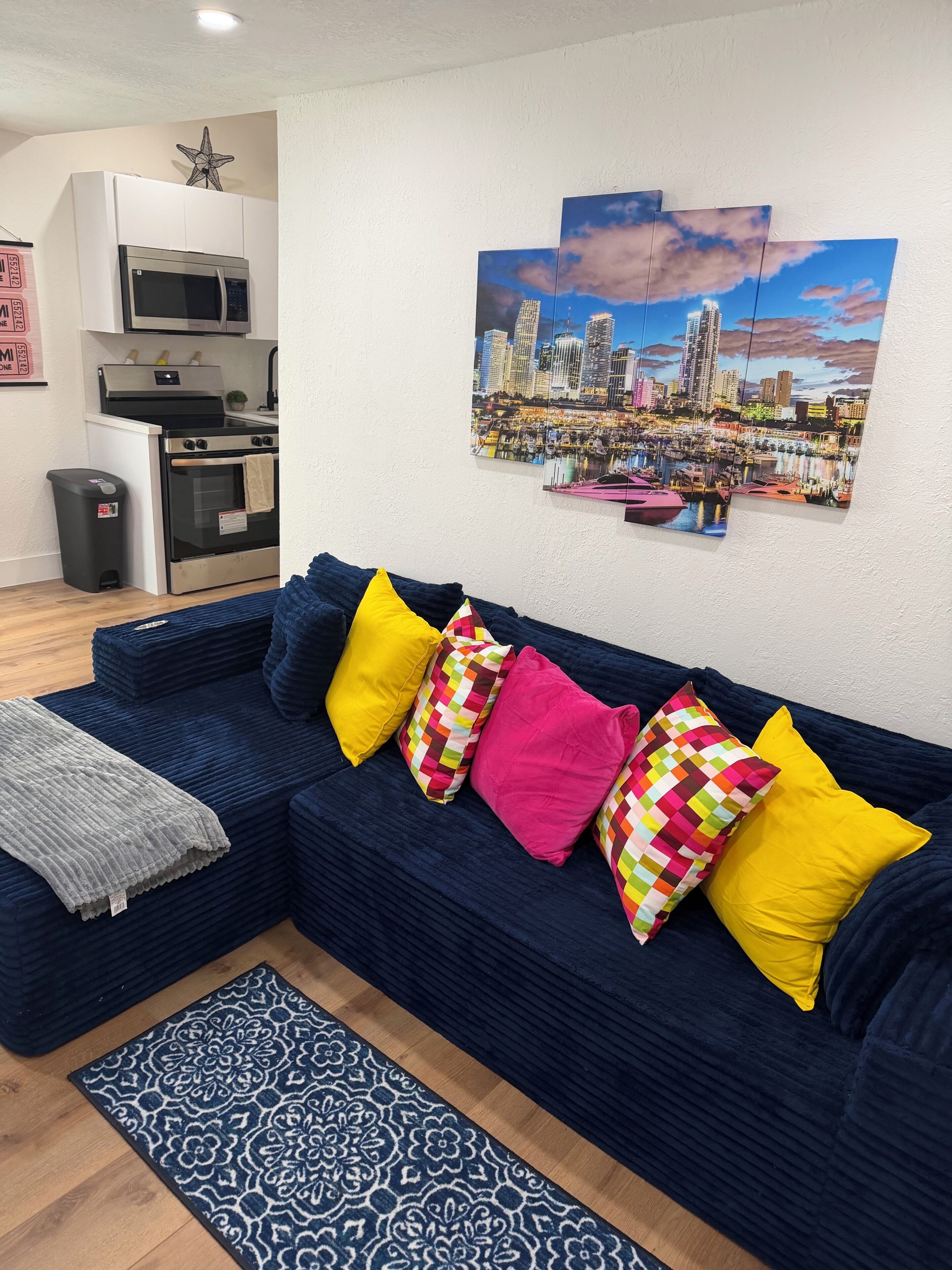 Ein Wohnzimmer mit einem dunkelblauen Sofa, bunten Kissen, einem gemusterten Teppich und einem Stadtbild an der Wand.