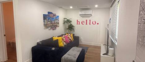 A living room with a dark blue sofa, a small table with a plant, and a wall-mounted air conditioner.