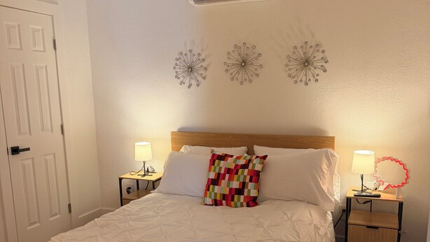 A neatly made bed with a quilted white duvet, a colorful pillow, and a wooden headboard. There is a wall-mounted air conditioner above the bed, and a small bedside table with a lamp and a phone. - Cozy 1-bedrm apartment with AC in vibrant Miami near Wynwood & Design District! (Miami)