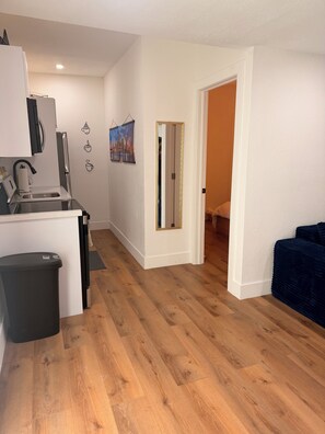 A modern kitchen with a sink, refrigerator, and a trash bin. There is a hallway leading to another room with a door and a blue couch. - Cozy 1-bedrm apartment with AC in vibrant Miami near Wynwood & Design District! (Miami)