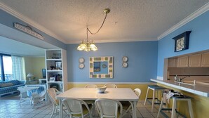 A dining area with a table and chairs, a kitchen with cabinets, a clock, and a sign on the wall.