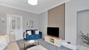 A modern living room with a sofa, a coffee table, a TV on a stand, a door, and a wall-mounted panel. - Spacious 3BR Hawick Apartment, Sleeps 6, Parking (Scottish Borders)