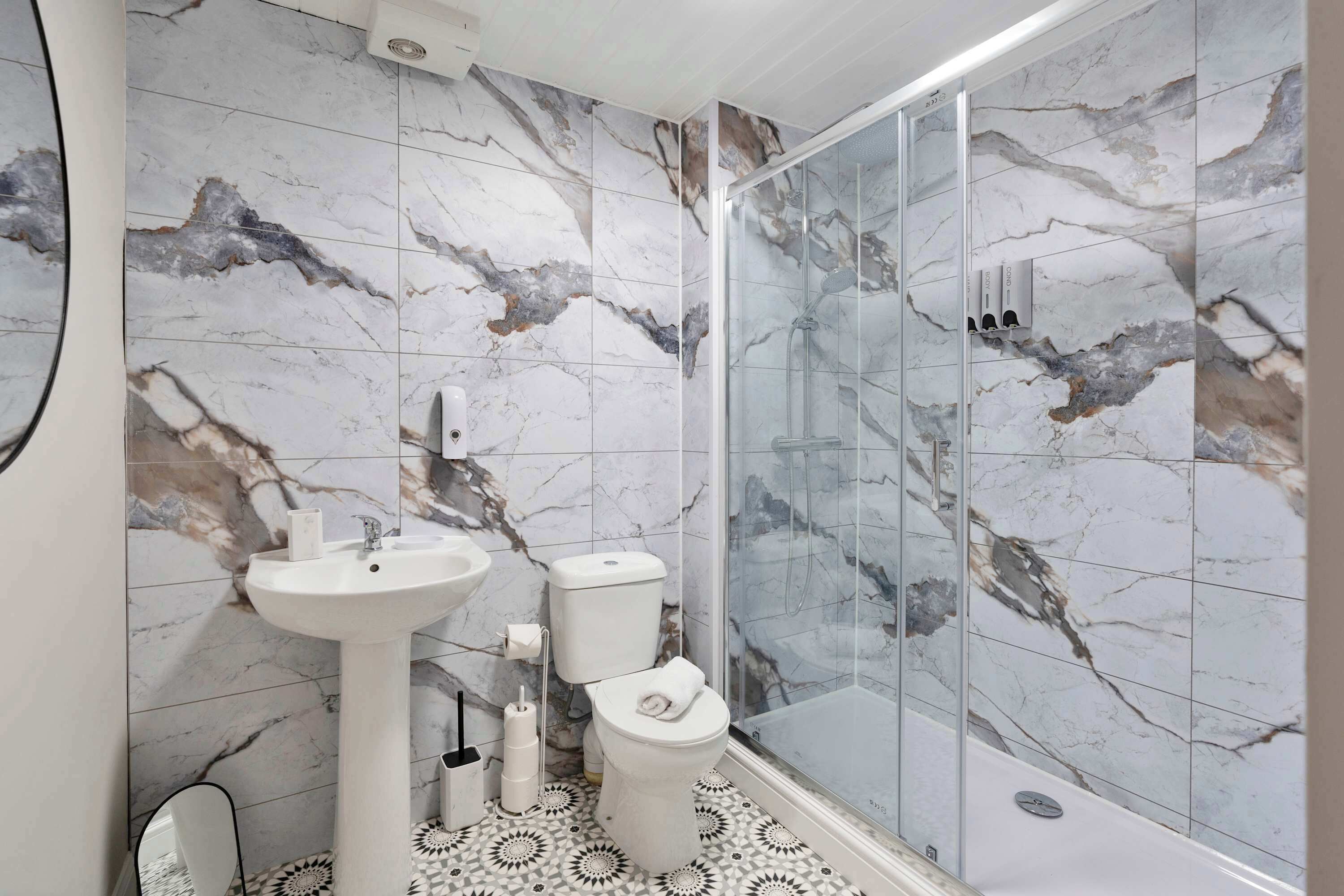 Et badeværelse med marmorbeklædning, et pedestalvask, et toilet og en brusekabine med glasvægge.