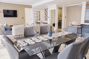 A modern living room with a glass dining table, grey sofas, and a TV mounted on the wall. - Newly Upgraded Stays (Sandton)