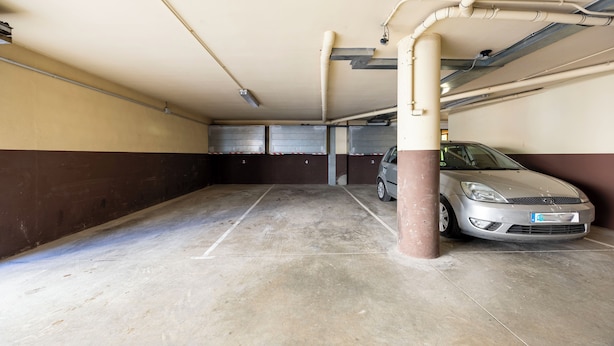 A parking garage with concrete floors and walls, a car parked in one of the spaces, and visible pipes and lighting fixtures. - La Molina: Ski-in apartment with private parking (Alp)