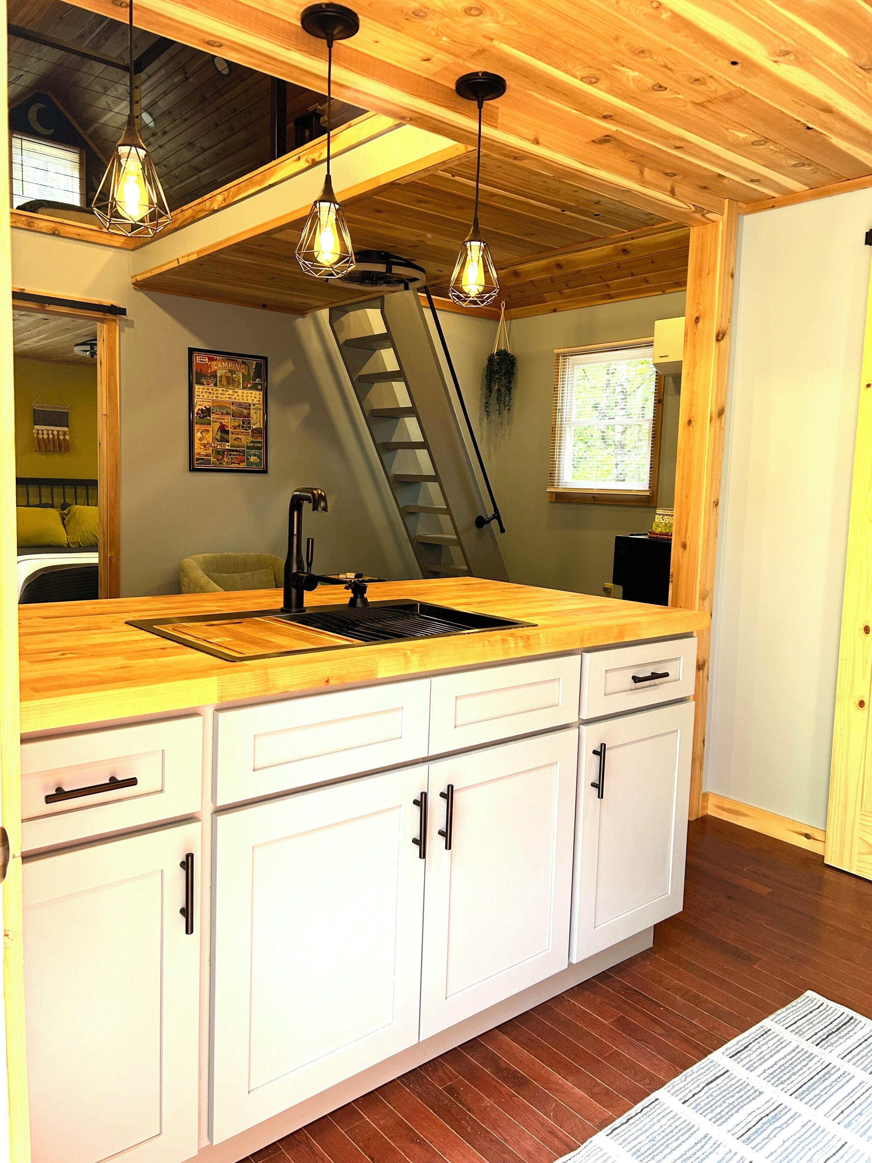 Une cuisine avec des meubles en bois, un îlot central, des luminaires suspendus, une fenêtre avec des stores et un escalier.
