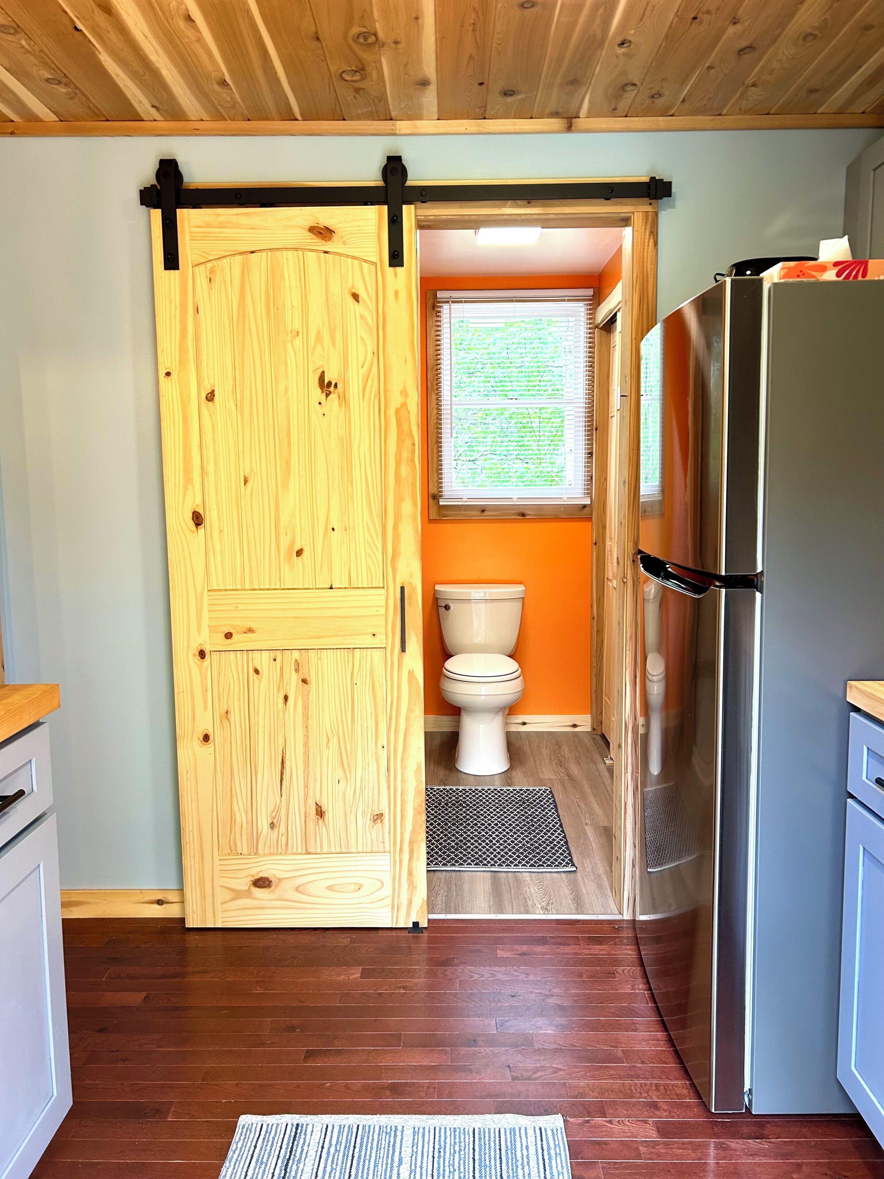 Un intérieur en bois avec une porte coulissante donnant accès à une salle de bain, un réfrigérateur en acier inoxydable et une fenêtre équipée de stores.