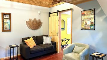 Un salon avec un canapé foncé, un fauteuil clair, un plafond en bois, un tableau encadré et une porte donnant sur une autre pièce.