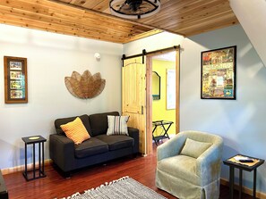 A living room with a dark sofa, a light armchair, a wooden ceiling, a framed picture, and a door leading to another room.