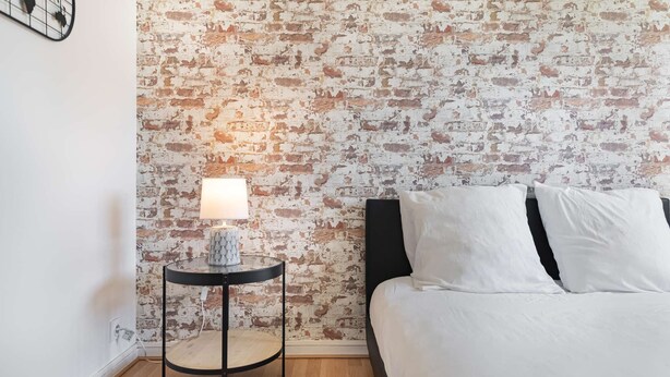 Un dormitorio con una pared de ladrillo, una mesa redonda con lámpara y una cama con ropa blanca.