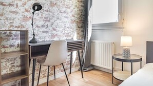 Una habitación con pared de ladrillo, un escritorio con silla, una lámpara y una mesita de noche con lámpara.